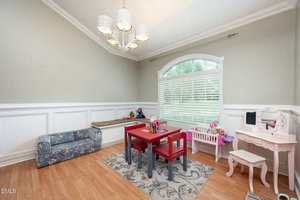 A children's playroom with a small red table and matching chairs on a rug, a floral-patterned sofa, toy vanity set, baby doll crib, and a large window with white blinds letting in natural light.