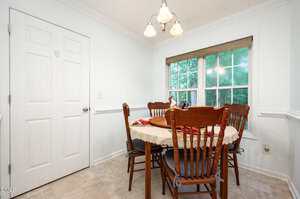 A small dining area features a round wooden table with a beige tablecloth, surrounded by four wooden chairs, positioned near a window with a view of greenery and next to a closed white door.