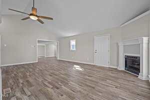 Spacious empty living room with light-colored walls, vaulted ceiling, wood-look flooring, a ceiling fan, a small window, white doors, and a fireplace with a decorative mantel in the corner.