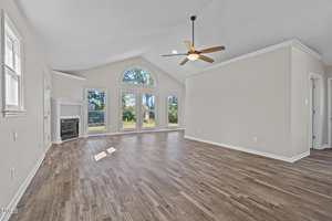 Spacious living room with vaulted ceiling, large arched window, several rectangular windows, ceiling fan, light gray walls, wood-look flooring, and a white fireplace in the corner.