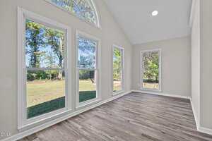 A bright room with large windows, light-colored walls, and wood-look flooring; the windows provide a view of trees and a grassy yard outside. The room has a vaulted ceiling with a circular window at the top.