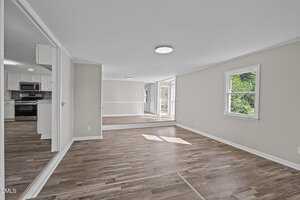 A spacious, empty room with wood flooring, white walls, and a window on the right. The room opens to a kitchen with white cabinets and stainless steel appliances, and a dining area with glass doors leading outside.