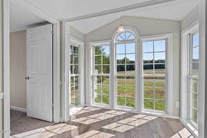 A bright sunroom with large, white-trimmed windows and a glass door, allowing natural light to fill the space. The room features light wood flooring and a view of a grassy field and trees outside.