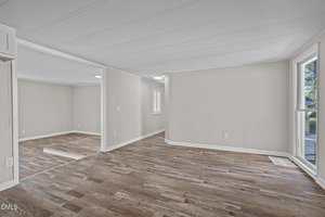 Empty room with light beige walls, white trim, and wood-look vinyl flooring. Large windows let in natural light. The space is open with visible doorways connecting multiple rooms.