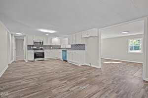 A spacious, open-concept kitchen and living area with wood-style flooring, white cabinets, stainless steel appliances, a blue dishwasher, and a window letting in natural light. A second room is visible through a wide doorway.