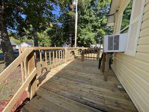 A wooden deck with railings and steps leads to a yellow house with white shutters. An air conditioning unit is mounted in the window. Trees and power lines are visible in the background.