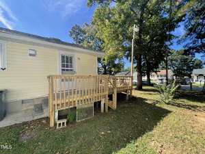 A wooden deck with railings is attached to the back of a yellow house. The yard has grass, several large trees, and a clear blue sky overhead. A street with houses and parked cars is visible in the background.
