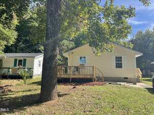 A small, single-story beige house with a wooden deck and white trim sits in a grassy yard with a large tree in the foreground and another white house visible to the left.