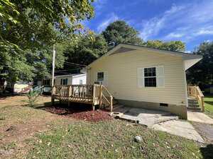 A single-story beige house with white trim and shutters, featuring a wooden accessibility ramp leading to the front door. The yard has grass, mulch, and trees, and it's a sunny day with a blue sky.