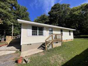 A single-story beige house with white trim sits on a grassy lawn at 215 Wilmington Avenue. Three front windows, a front door with a small wooden porch and steps, and trees in the background complete this Oxford, NC 27565 home.