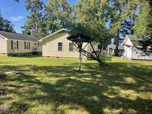 A grassy backyard with three light-colored single-story houses, several trees providing shade, a birdhouse mounted on a wooden post, and a satellite dish on the right side near one house.
