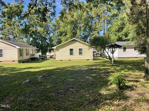 A grassy backyard with three single-story beige houses, surrounded by trees. One house is centered, another is on the left, and a third is partially visible on the right with a small black-roofed structure nearby.