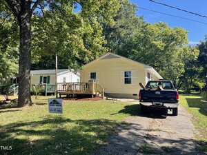 A light yellow single-story house with a wooden front deck sits beside a white house. A black pickup truck is parked in the driveway. A yard sign for a real estate agency is posted on the lawn under a large tree.