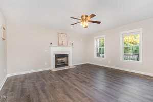 A spacious living room with light beige walls, dark wood flooring, a white fireplace with art above it, a ceiling fan with lights, and two large windows letting in natural light.