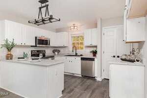 A modern kitchen with white cabinets, stainless steel appliances, light gray walls, marble countertops, a large center island, and wood-look flooring. Decorative plants and dishes are placed on the counters.