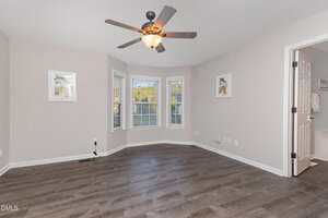 Unfurnished room with gray walls, dark wood flooring, a ceiling fan with light, bay windows with white blinds, wall art, visible outlets, and an open door leading to a bathroom.