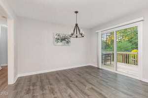 A dining area with light gray walls, dark wood flooring, a black chandelier, wall art, and large sliding glass doors leading to a wooden deck overlooking a green, wooded area.