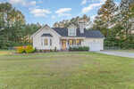 A white, single-family house at 1701 Cobblestone Drive in Creedmoor features a covered front porch with an American flag, attached garage, manicured lawn, and is surrounded by trees under a blue sky with scattered clouds.