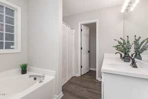 A bathroom with a white bathtub, a double sink vanity with two mirrors, a vase with greenery, a shower with a curtain, and a door. The walls are light-colored and the floor has dark wood-style flooring.
