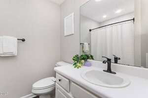 A bathroom with a white sink, black faucet, white countertop, and cabinet. There is a toilet, a towel on a black rack, a shower with a white curtain, and a mirror above the sink. A plant and soap are on the counter.
