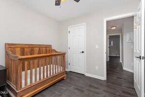A nursery room with a wooden crib, white walls, dark wood flooring, and two white doors; one door is open, revealing a hallway with a light fixture and framed artwork on the wall.