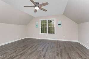 An empty room with light-colored walls, a window centered on the far wall, two small pieces of abstract art, dark wood flooring, and a ceiling fan with wooden blades.