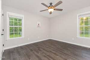 A bright, empty room with light gray walls, two large windows with white trim, dark wood flooring, a ceiling fan with a light, and one framed picture on the wall. Greenery is visible outside the windows.