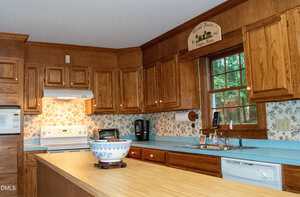 A kitchen with wood cabinets, a light blue countertop, floral wallpaper, and a large window over the sink. Appliances include a white stove with range hood, microwave, dishwasher, and a blue-and-white bowl on the island.