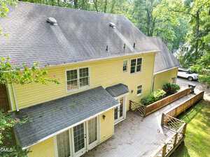 Aerial view of the back of a two-story yellow house with a gray shingled roof, a large wooden deck, multiple windows, and a driveway leading to a white parked vehicle surrounded by trees.