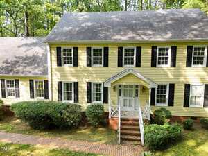 Two-story yellow house with black shutters, white trim, and a covered front porch with stairs. Brick walkway leads to the entrance. Shrubs and grass surround the house, and trees are visible in the background.