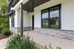 Front porch of a modern house with white vertical siding, stone accents along the lower wall, black-framed windows, a concrete walkway, and landscaped bushes in the foreground.