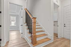 A hallway in a modern home features wooden stairs with black railings, light-colored walls, and light wood flooring. Multiple doors lead to adjacent rooms, including a laundry area and a room with a window.