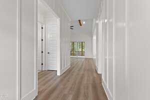 A bright hallway with white paneled walls and wood flooring leads to a living area with large windows and glass doors opening to a balcony or porch. Exposed wooden beams are visible on the ceiling in the distance.