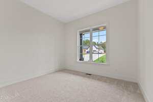 A small, empty room with beige carpet, white walls, and a large window showing a view of a residential street and neighboring houses outside.