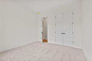 Empty room with light beige carpet, white walls, a closed double-door closet, and an open door leading to a hallway with wooden flooring. The room has simple baseboards and no furniture or decor.
