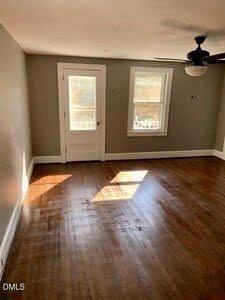 An empty room with hardwood floors, beige walls, a white door leading outside, two windows, a ceiling fan with a light, and sunlight streaming onto the floor.