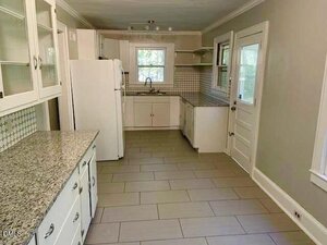 A galley kitchen with white cabinets, granite countertops, tile flooring, a white refrigerator, sink under a window, tiled backsplash, and a door leading outside. There are upper and lower cabinets for storage.
