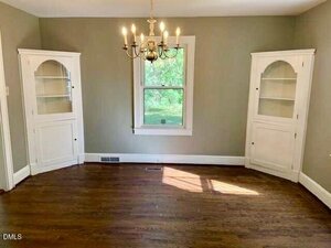 A dining room with hardwood floors, a window centered on the back wall, a brass chandelier, and two white built-in corner cabinets with shelves and closed storage at the bottom.