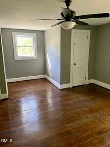 An empty room with gray walls, a white ceiling, a window, a white door, a ceiling fan with light, and hardwood floors. Natural light is coming in through the window.