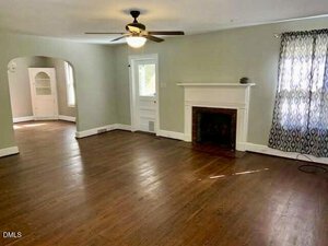 A spacious living room with hardwood floors, a ceiling fan, a white fireplace, light green walls, and a window with patterned curtains. An arched doorway leads to an adjacent room with built-in shelves.