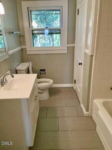 A small bathroom with a white vanity and sink, a toilet, a window with trees visible outside, beige tiled floor, and a bathtub with white tiles on the right. A towel bar is mounted on the left wall.