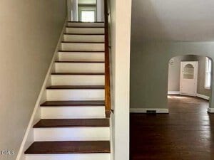 A staircase with wooden steps and white risers is on the left, leading to an upper floor. To the right is a room with hardwood floors, an arched doorway, and a built-in white cabinet near a window.