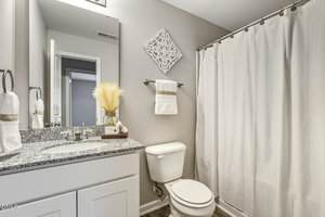 A small bathroom with a white vanity and granite countertop, a toilet, a shower with a white curtain, neutral wall decor, and towels neatly arranged. The room has light grey walls and modern fixtures.