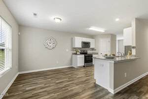 Modern kitchen and dining area with wood-style flooring, white cabinets, stainless steel appliances, granite countertops, and a decorative wall clock. Large window provides natural light.