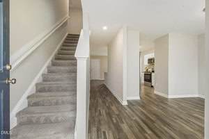 A carpeted staircase is on the left side of an entryway with wood-style flooring. A hallway leads to a kitchen with white cabinets and stainless steel appliances. Walls are painted light beige.