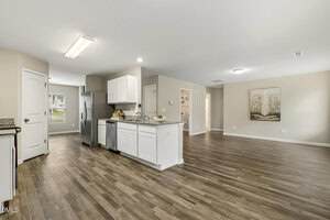 Open-concept kitchen and living area with white cabinets, stainless steel appliances, granite countertops, light gray walls, wood-look flooring, a window, and a doorway leading to another room.
