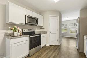 Modern kitchen with white cabinets, stainless steel appliances, granite countertops, a vase of flowers, and a tray with cups on the counter. A pantry door and a bright dining area with windows are visible in the background.