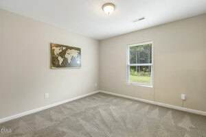 A simple, empty room with beige walls and carpet, one window with blinds, a ceiling light fixture, and a world map artwork hanging on the wall. Sunlight is coming in through the window.