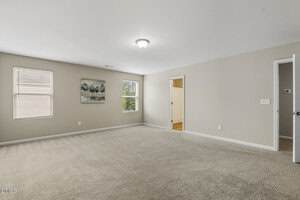 A spacious, empty room with beige walls and carpet, two windows with blinds, a ceiling light fixture, a wall painting, and two open doorways leading to other rooms.