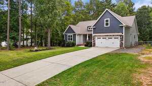A two-story house with dark gray siding and a two-car garage sits at the end of a long concrete driveway, surrounded by a lawn and tall trees in a wooded neighborhood.
